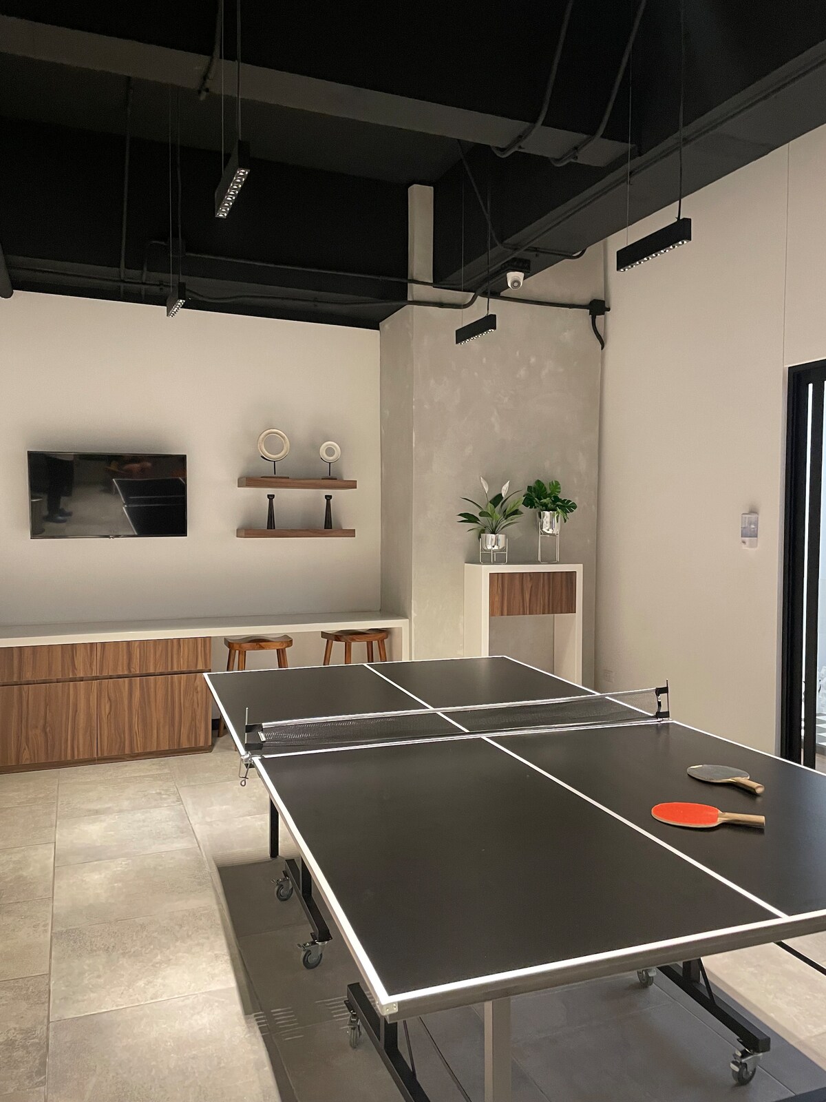 A recreational area is equipped with a ping pong table at the center. Modern stools are positioned nearby. A wall-mounted television and minimalistic shelves with decorative items are visible in the background, contributing to a relaxed entertainment space.