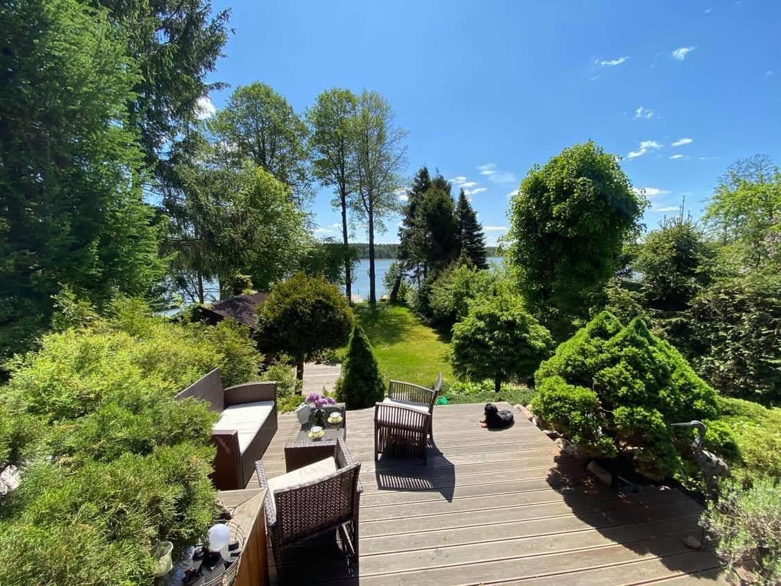 A spacious outdoor deck is surrounded by lush greenery and offers a view of the lake. Comfortable seating arrangements are positioned for relaxation, with small potted plants adding to the natural ambiance. Tall trees provide shade, contributing to a serene atmosphere.