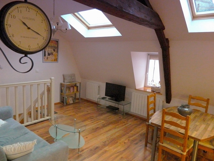 An open living area features wooden beams and plenty of natural light from skylights. A comfortable couch is positioned near a glass table, while a wooden dining table is set for four. A flat-screen television is placed on a glass stand, with bookshelves adding charm.