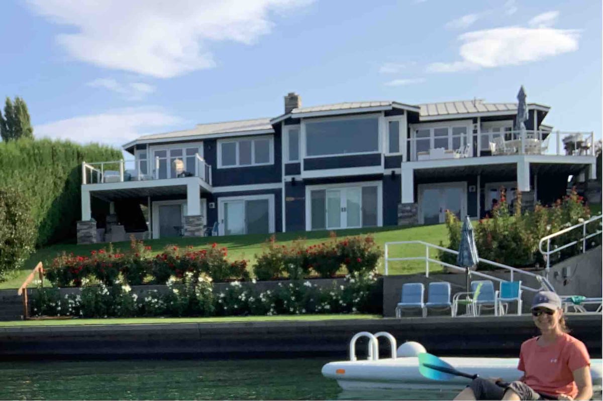 The lakefront home features a modern two-story design with large windows and multiple balconies overlooking the water. A spacious lawn adorned with colorful flowers leads to the lake. Comfortable outdoor seating is positioned on the lower deck, inviting relaxation by the water.