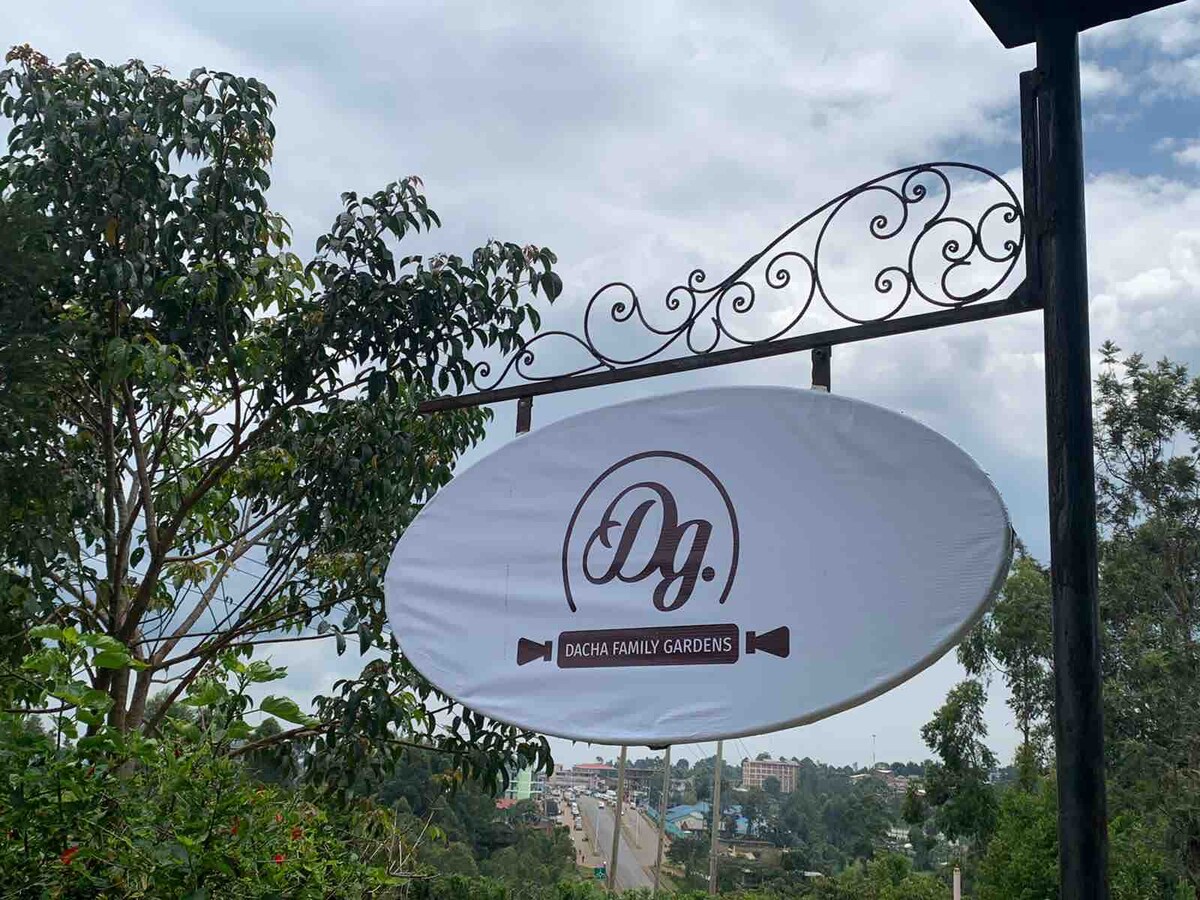 A sign for Dacha Family Gardens is displayed prominently, featuring the logo in elegant lettering. Surrounding foliage frames the sign, providing a natural element. In the background, a glimpse of the road is visible, suggesting accessibility to the location.
