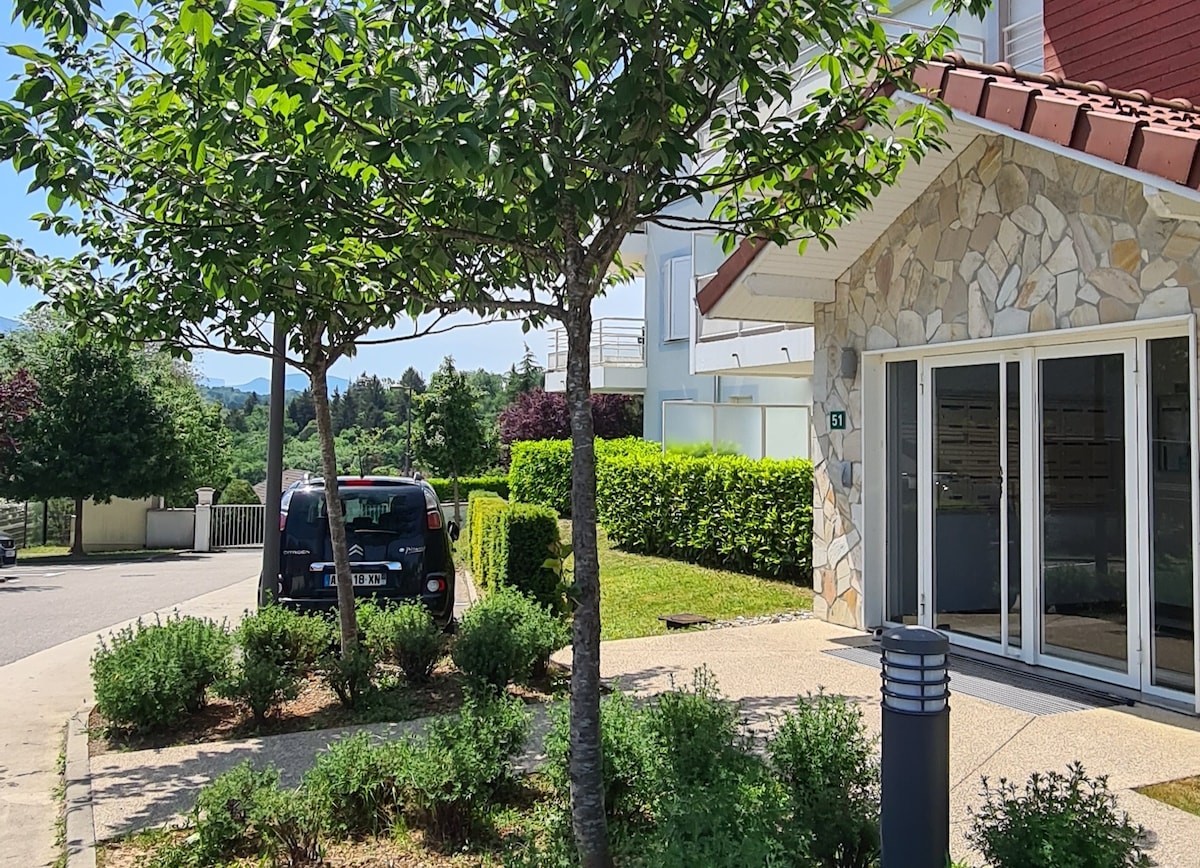 A modern property is showcased with natural stone accents and large glass doors. Surrounding greenery includes well-maintained shrubs and trees, adding a touch of nature. A parked vehicle is seen nearby, indicating convenient access and a welcoming approach to the entrance.
