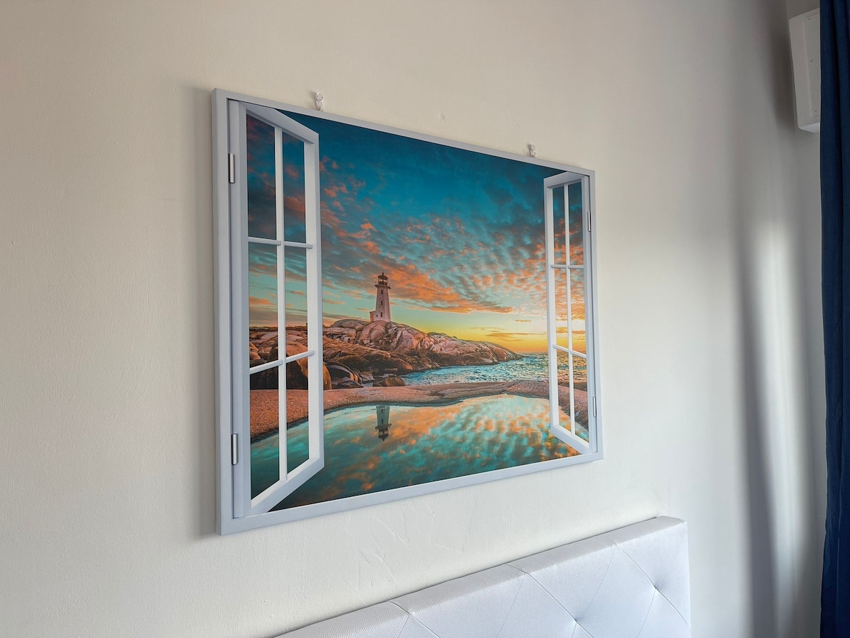 A large wall art piece depicting a serene coastal scene is displayed above the bed. The artwork features a lighthouse against a vibrant sky at sunset, framed by an open window. Soft colors and reflections of water are highlighted, creating a calming visual.