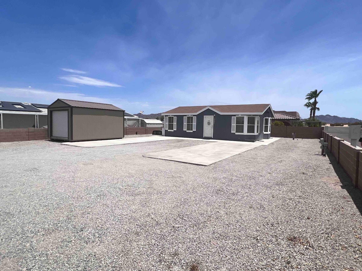 A modern one-bedroom home is visible on a spacious gravel lot, accompanied by a storage shed. The house features multiple windows framed in white, and a front porch with a simple entrance. Surrounding areas indicate ample parking space suitable for larger vehicles.