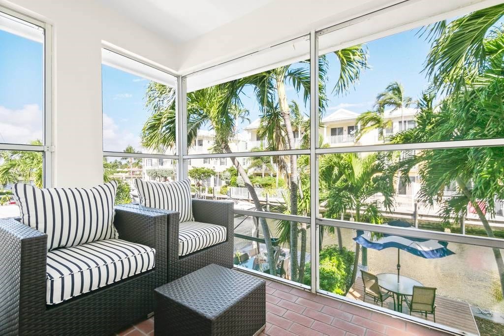 A bright and airy seating area is furnished with two striped chairs and a matching coffee table. Large windows offer views of lush palm trees and the waterfront canal, creating a relaxing environment for indoor relaxation.
