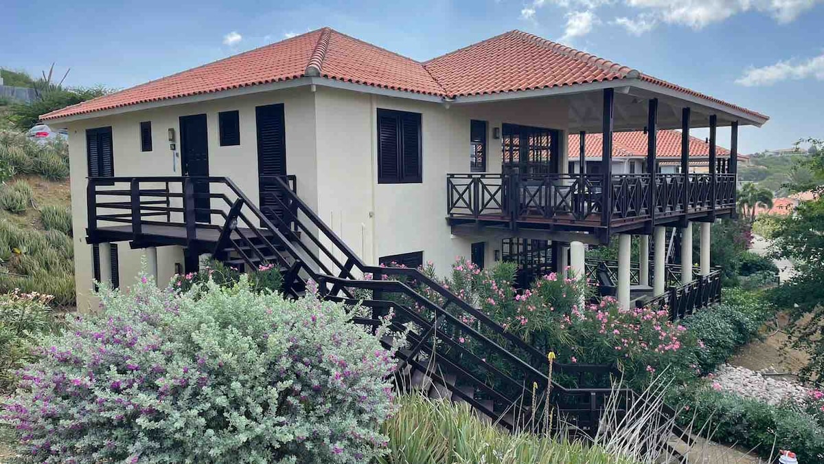 A two-story villa features a terracotta-tiled roof and wooden railings. Surrounded by lush greenery and flowering plants, the structure is elevated, providing a spacious outdoor area with multiple steps leading to the entrance. Large windows create a bright, inviting atmosphere.