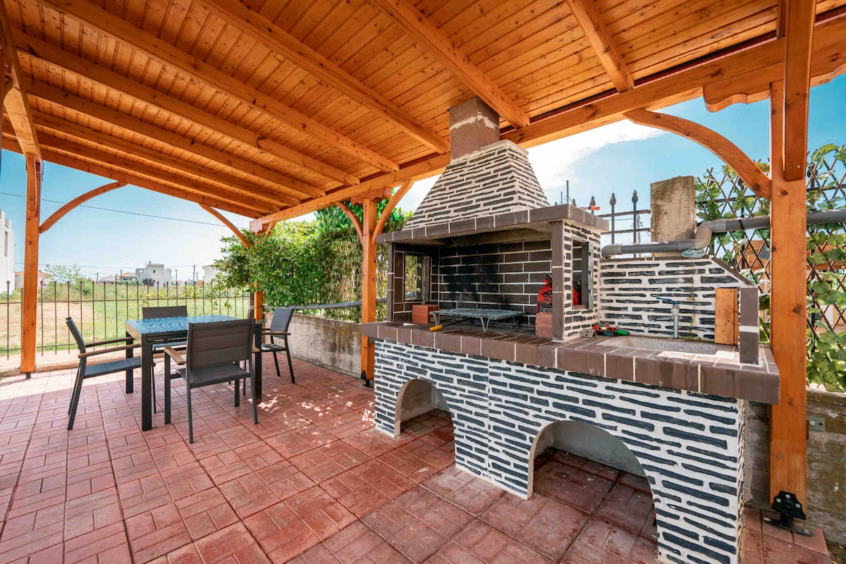 An outdoor patio features a covered seating area and a built-in stone BBQ grill. The space is surrounded by greenery and has a tiled floor, ideal for family gatherings or alfresco dining.
