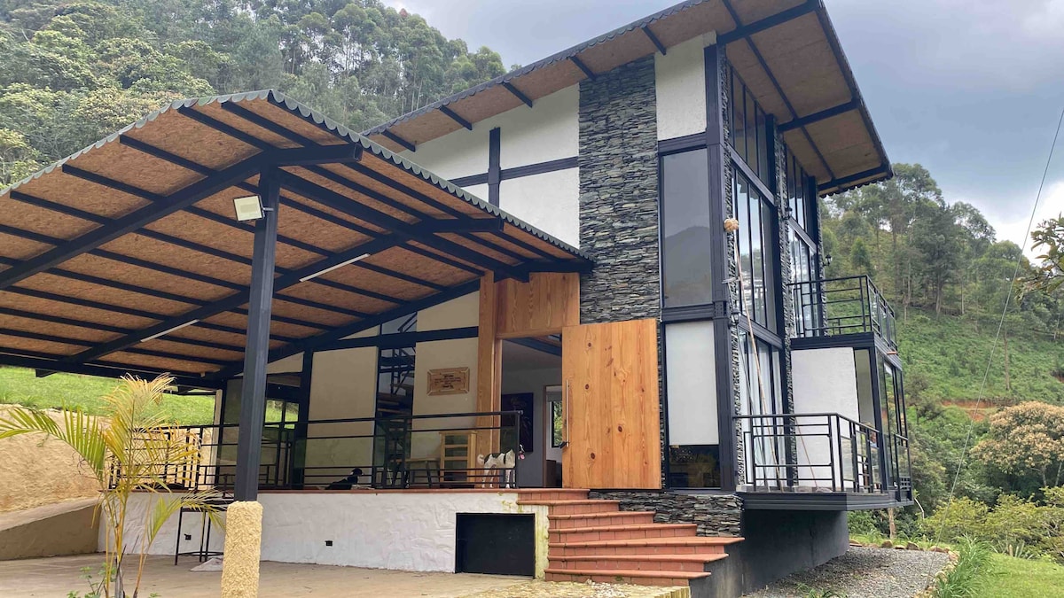The spacious cabin features a contemporary design with large windows that invite natural light. A blend of stone and wood elements enhances the exterior. A covered terrace with seating overlooks the surrounding lush greenery, providing an ideal space for relaxation.