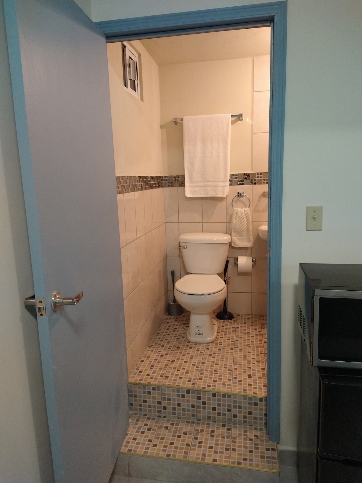 A compact bathroom is shown, featuring light-colored tiled walls and a mosaic tiled floor. A white toilet is positioned in the center, with a towel rack holding a neatly hung towel. A window is visible above, providing natural light.