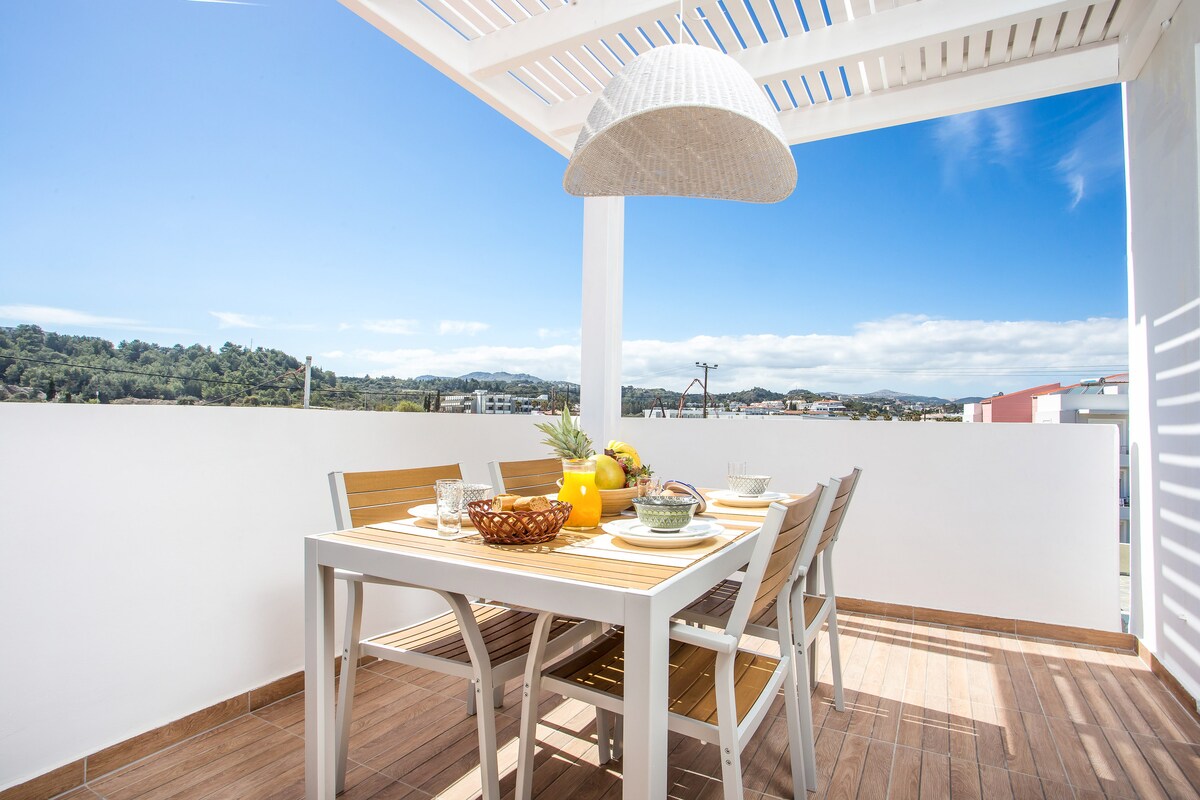 A dining area is presented on a sunlit balcony, featuring a table surrounded by six wooden chairs. A large pendant light hangs above, and the table is adorned with ceramic dishware, a bowl of fruit, and decorative items. Lush green hills are visible in the distance under a clear blue sky.