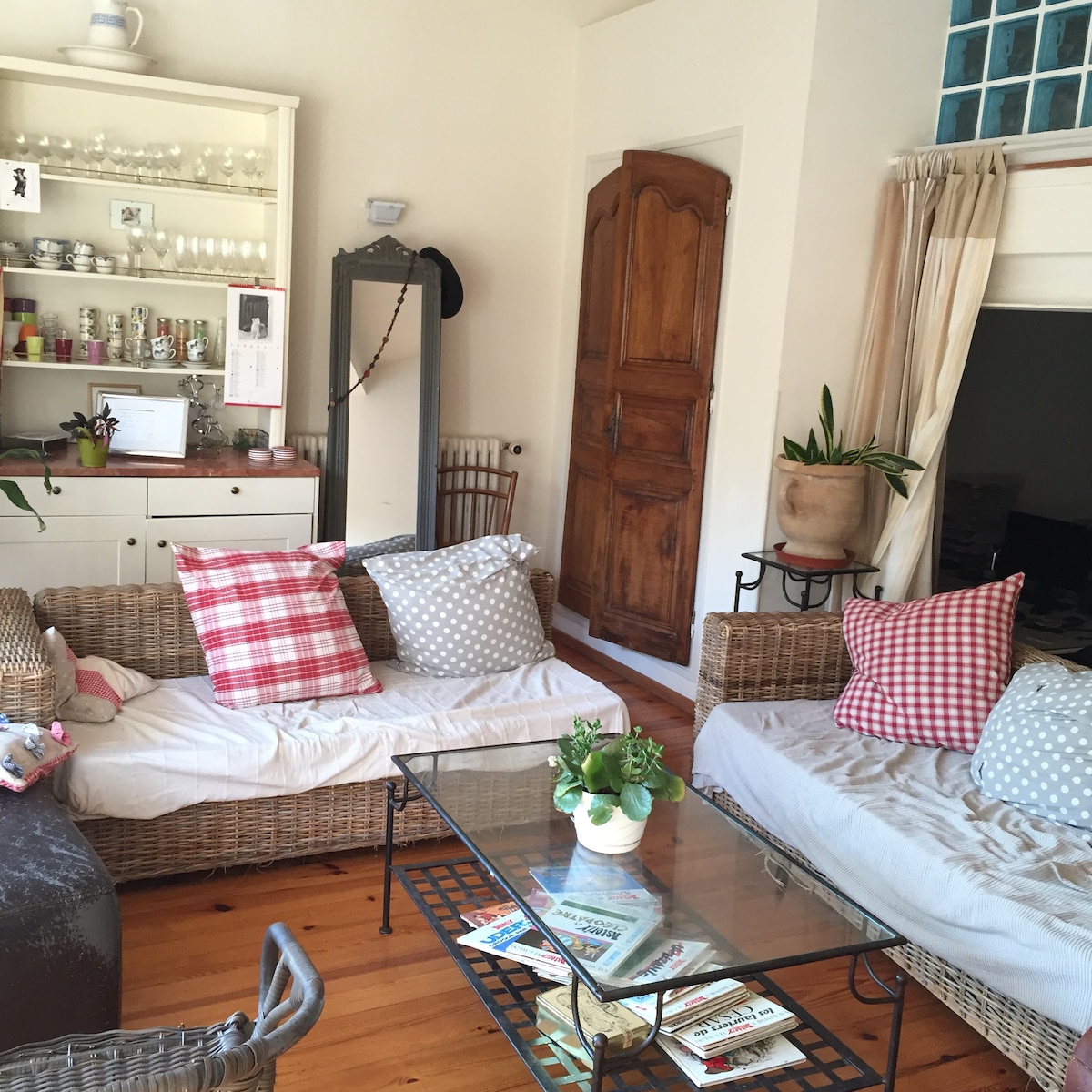 A living area features two rattan sofas with decorative cushions in various patterns. A glass coffee table is centrally placed, displaying magazines and a small potted plant. Natural light enters through a nearby window, enhancing the room's spaciousness.