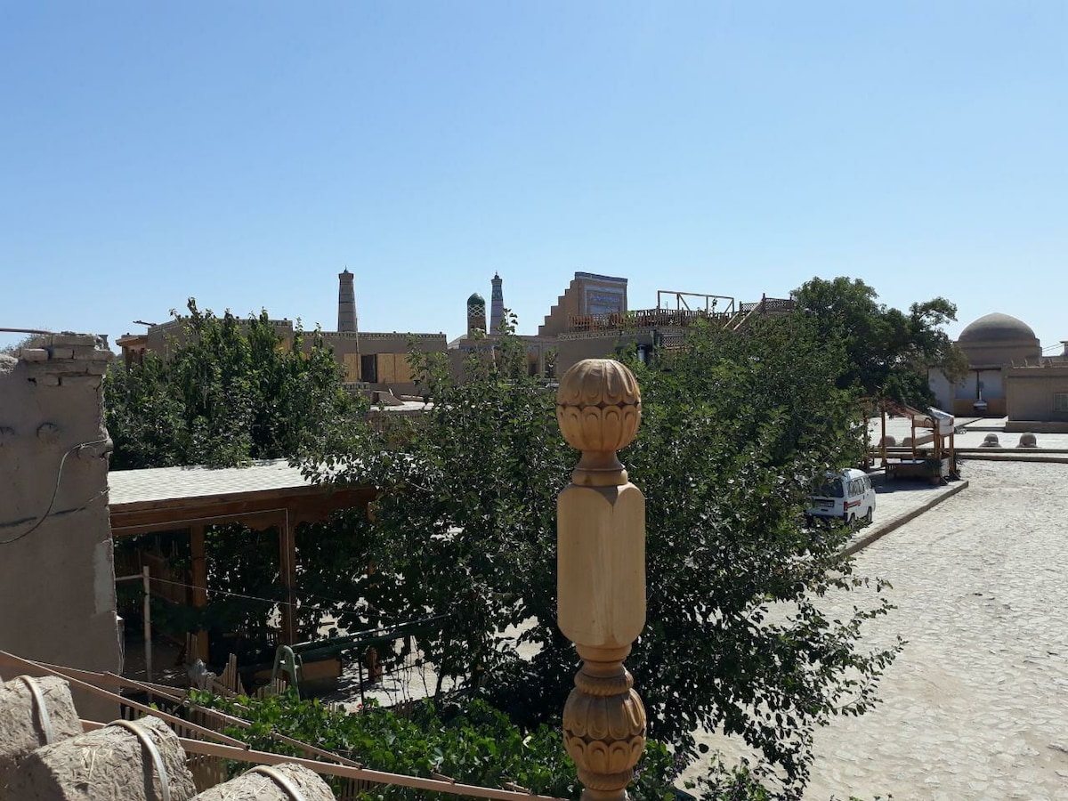 A view of a courtyard showcasing traditional architecture with tall structures in the background. Lush trees provide greenery, and a gravel pathway is visible. An open wooden structure offers shaded seating. Clear skies enhance the serene atmosphere of the surroundings.