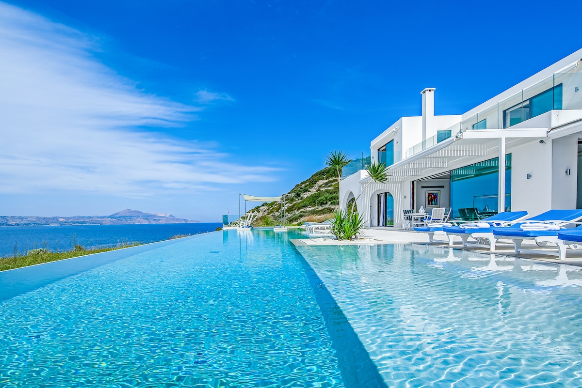 An infinity pool stretches towards the horizon, blending seamlessly with the shimmering sea. Modern lounge chairs are positioned around the pool, with the villa's contemporary architecture visible in the background. Clear blue skies and lush greenery complement the tranquil setting.
