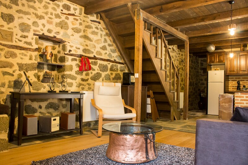 A warm living space features exposed stone walls and wooden beams, creating an inviting atmosphere. A comfortable chair is positioned next to a sleek coffee table. A staircase leads to the upper level, with kitchen amenities clearly visible in the background.