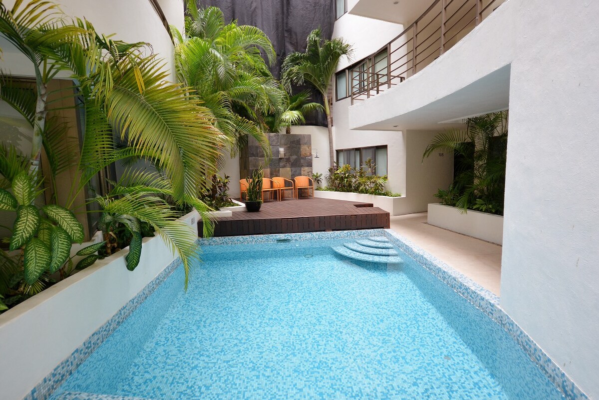 A sparkling swimming pool is surrounded by lush greenery and tropical plants, creating a serene atmosphere. Comfortable seating is placed adjacent to the pool area, with a wooden deck extending into the water, inviting relaxation in a tranquil environment.