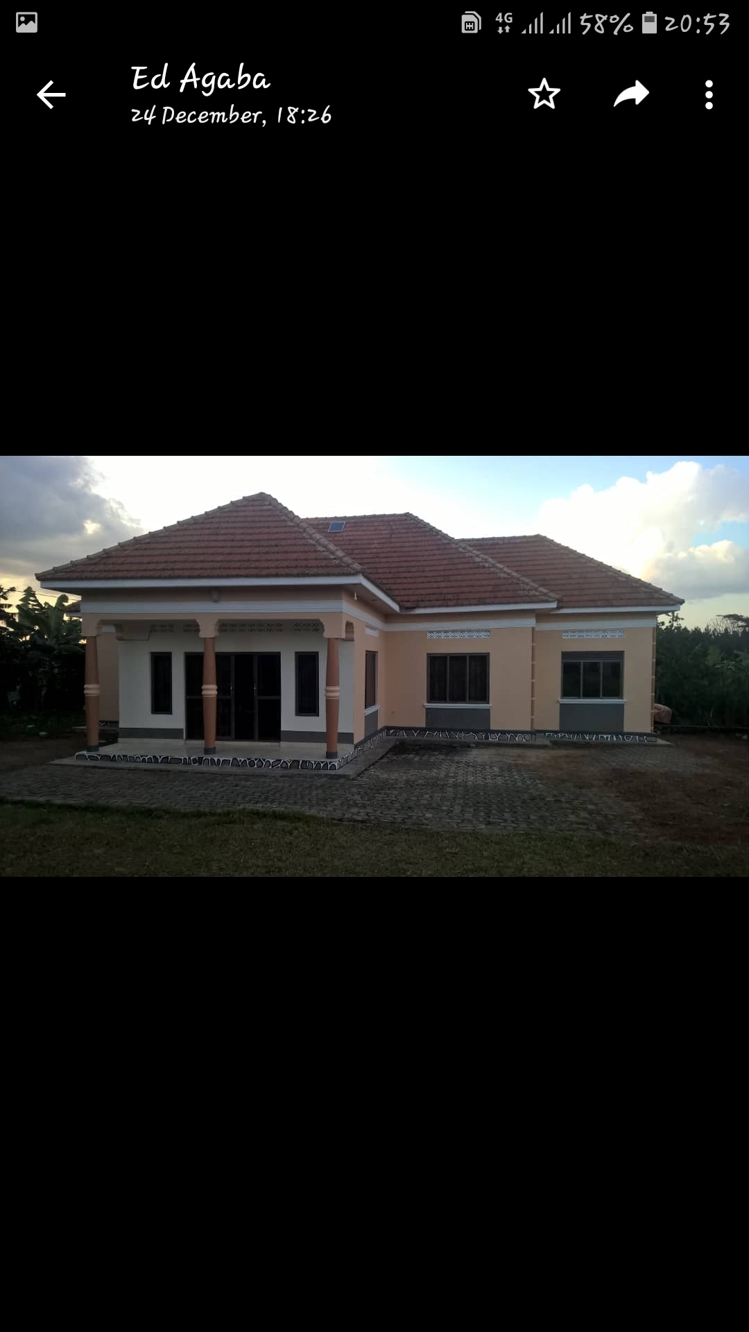 The home features a modern design with a sloped roof and large windows. A paved area surrounds the house, leading to a spacious entry. Lush greenery is visible in the background, enhancing the serene environment.