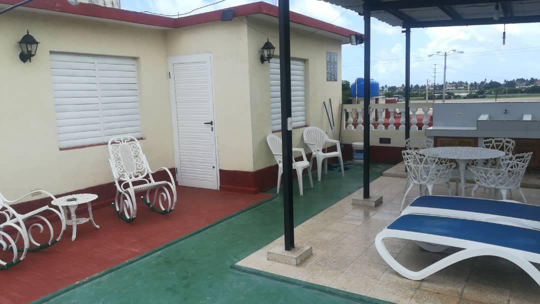 An outdoor terrace is featured with several white lounge chairs and tables set on a tiled floor. A shaded area contains a dining table surrounded by white chairs, with views extending to the surrounding area. Sun loungers invite relaxation under the open sky.