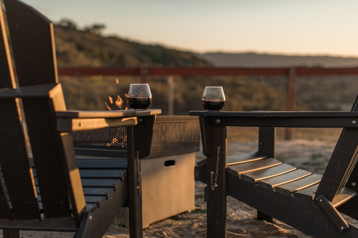 Two black Adirondack chairs are positioned near a small fire pit, with two glasses of red wine placed on the armrests. A scenic view of rolling hills is visible in the background, illuminated by the warm glow of the setting sun.