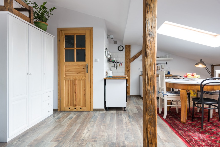 A bright and open living space features wooden beams and a large window with natural light streaming in. A round dining table is surrounded by chairs, with a fruit bowl as a centerpiece. A wooden door leads to an adjacent area, and storage is provided by a white cabinet.