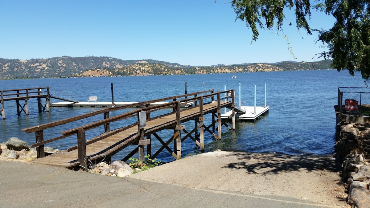 A view of a tranquil lake surrounded by rolling hills. Multiple wooden docks extend into the water, providing easy access for boaters. The clear blue sky reflects on the surface of the lake, creating a serene atmosphere. Vegetation borders the scene, enhancing the natural beauty.