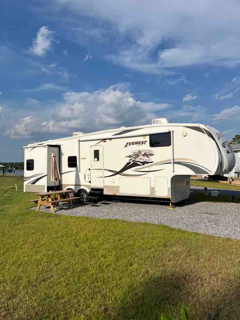 The camper, identified as Everest by Keystone, is parked on a grassy area. It features a large exterior with a retractable slide-out section. A small picnic table is positioned nearby, surrounded by well-maintained grass and gravel.