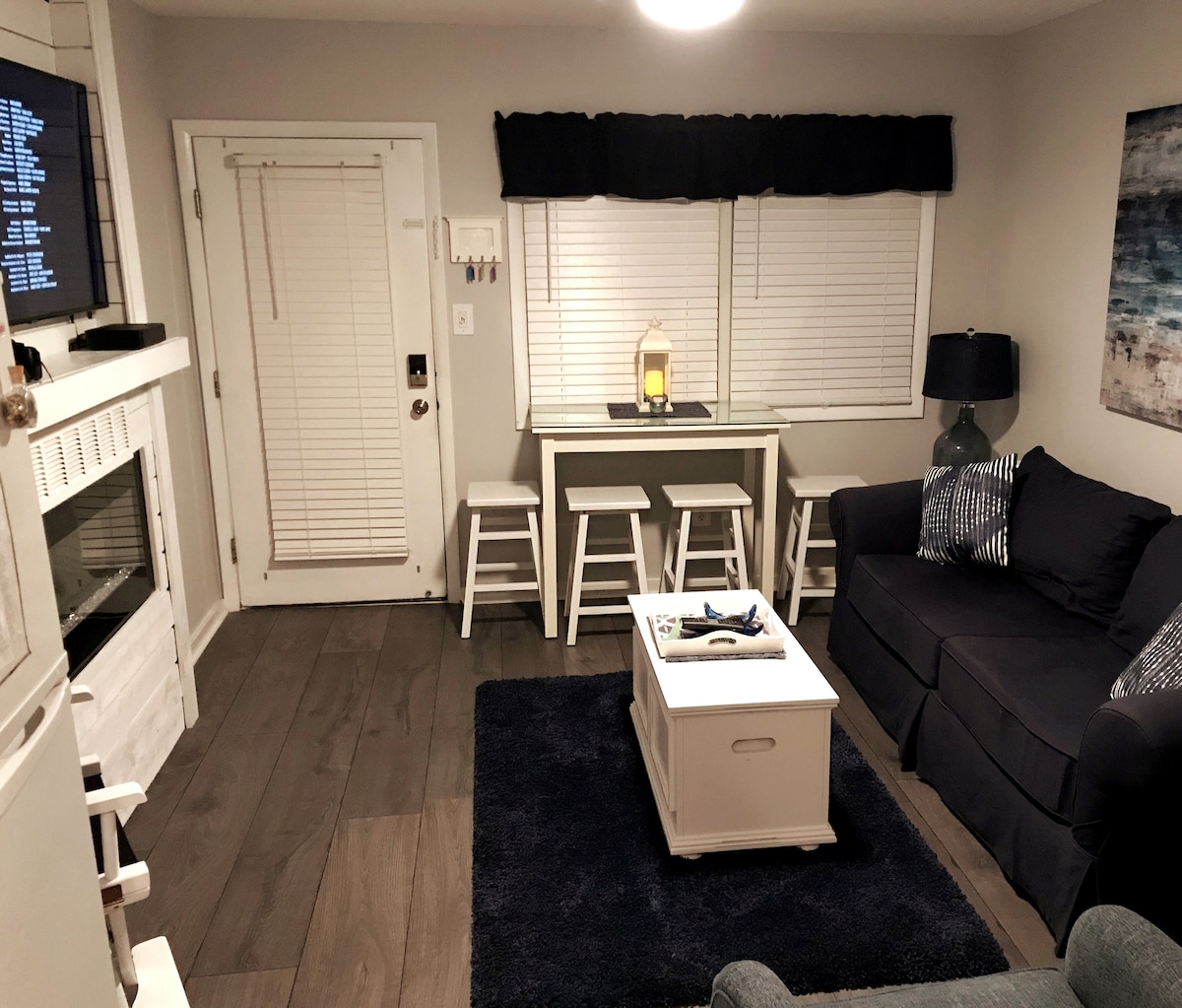 The living area features a cozy seating arrangement with a navy sofa and a light blue accent chair. A white coffee table is centered on a plush dark rug. A dining space with four white stools is positioned near a window, and a warm glow radiates from the lantern.