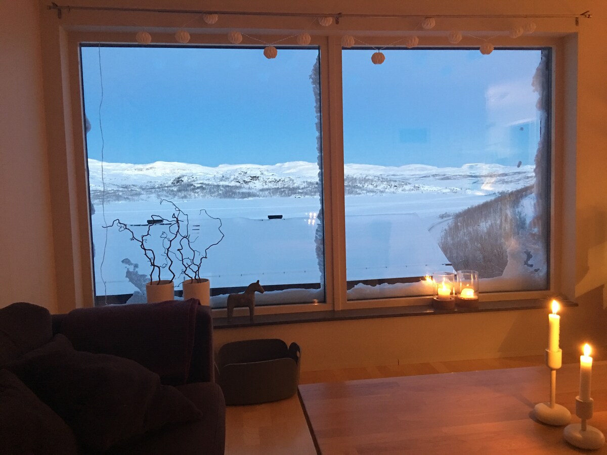 Large windows frame a serene winter landscape, revealing snow-covered hills and a gentle blue sky. The interior features a wooden table with lit candles and a cozy seating area, offering a warm and inviting setting to enjoy the view.