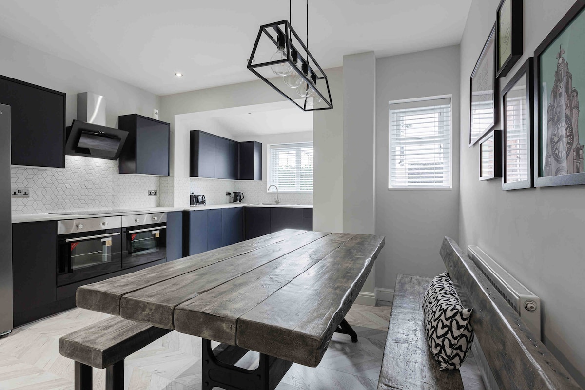 A modern kitchen is featured with dark cabinetry and stainless steel appliances. A spacious wooden dining table is centrally located, surrounded by a bench. Natural light enters through multiple windows, illuminating the room's minimalistic design.