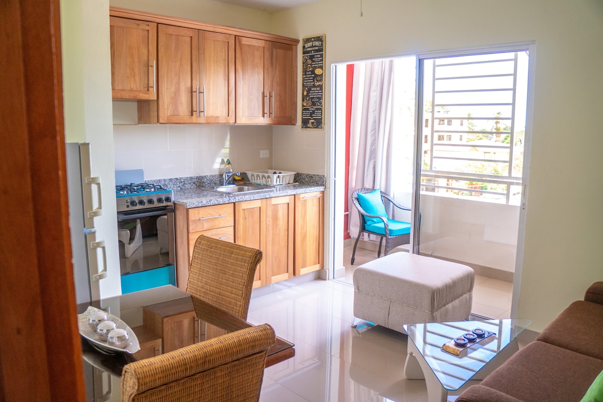 A well-organized kitchen area features wooden cabinetry and a granite countertop. Nearby, a cozy seating area consists of a soft ottoman, a glass coffee table, and a single chair. Natural light streams through the window, enhancing the open and airy ambiance.