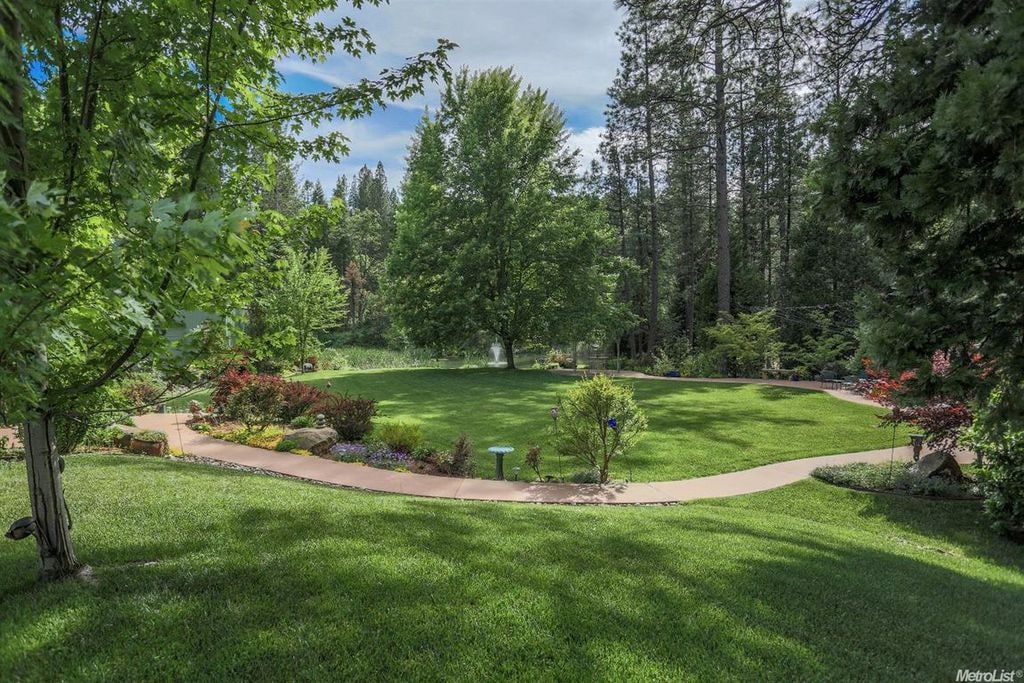 A lush green lawn is surrounded by trees and colorful plantings, creating a serene outdoor area. A pathway meanders through the landscape, leading towards a gentle pond and waterfall, inviting relaxation and exploration amidst nature.