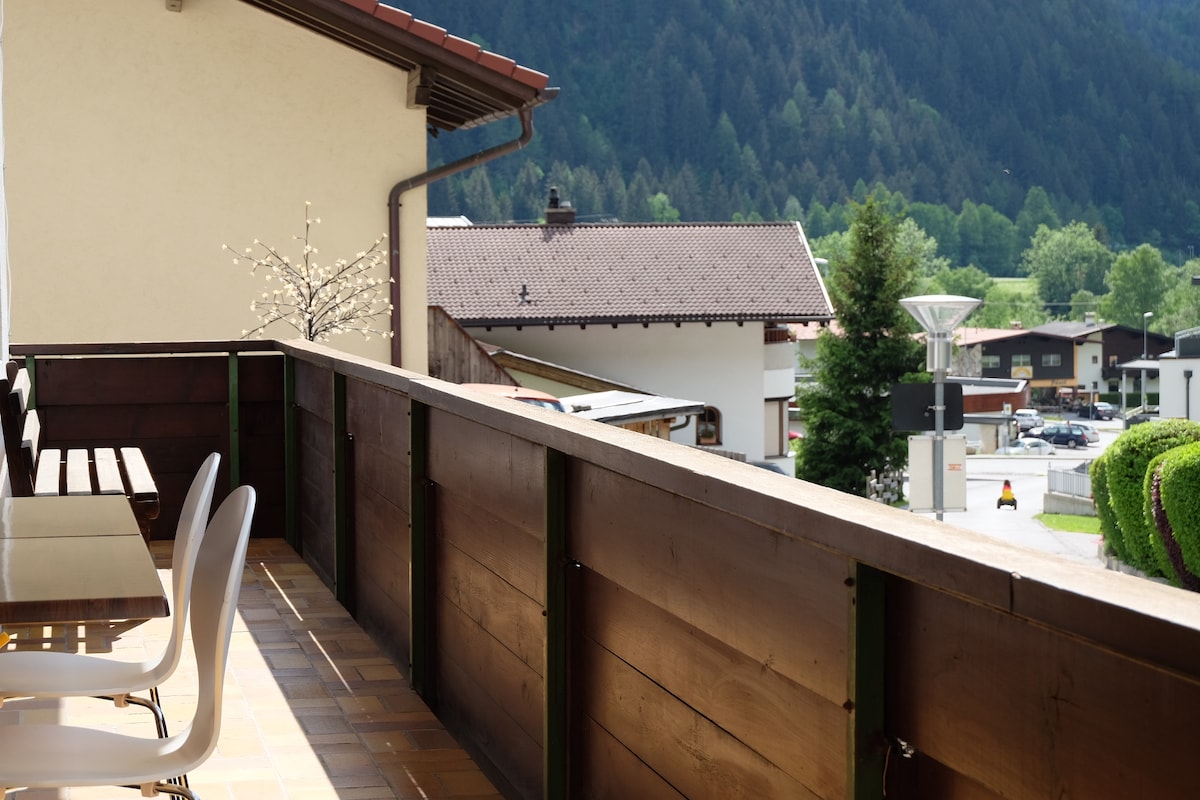 A private balcony is featured, equipped with a small seating area and table positioned against a wooden railing. Nearby views of surrounding green trees and residential structures are visible, with mountains rising in the background. Sunlight reflects off the surface, creating a bright atmosphere.