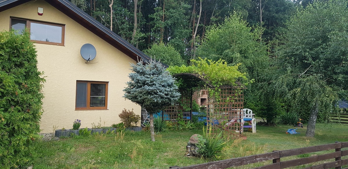 A charming house is surrounded by a lush garden featuring various trees and plants. A shaded patio area with a lattice trellis is visible, along with a brick grill. An inviting seating area is present, enhancing the outdoor space for relaxation.
