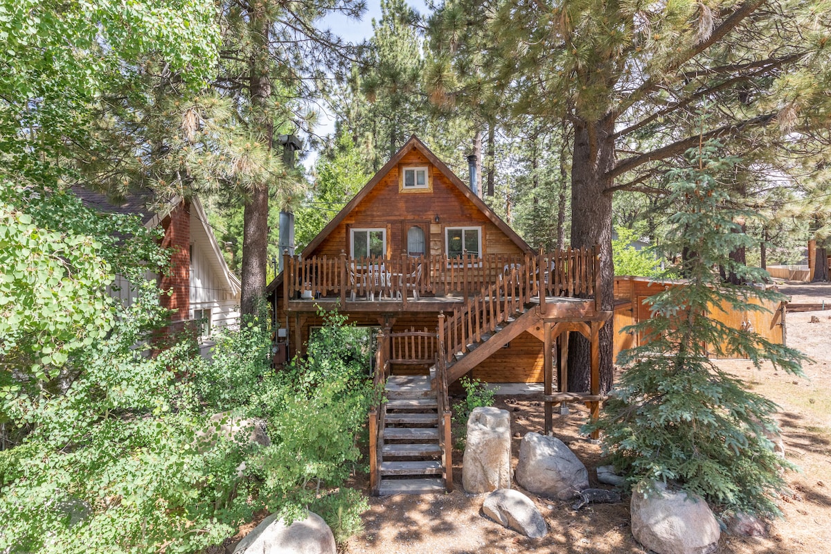 The cabin is surrounded by tall trees, with a welcoming wooden exterior and a spacious deck. A set of stairs leads to the entrance, flanked by rocks and greenery, highlighting the natural setting of the mountain town.