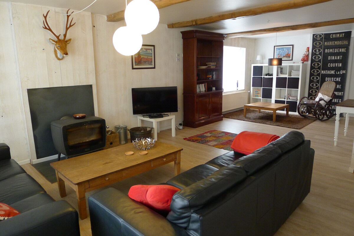 A spacious living room features comfortable leather sofas arranged around a wooden coffee table. A wood stove provides warmth, while a television and entertainment unit are visible. Natural light streams in through a window, illuminating the neutral decor and wooden beams on the ceiling.