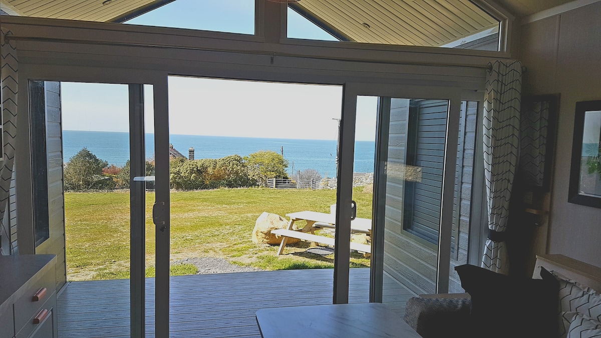 Large sliding glass doors provide a panoramic view of the sea and surrounding landscape. The open space leads to a deck area featuring outdoor seating. A grassy area extends toward the horizon, where the sky meets the water, creating a calm and serene atmosphere.
