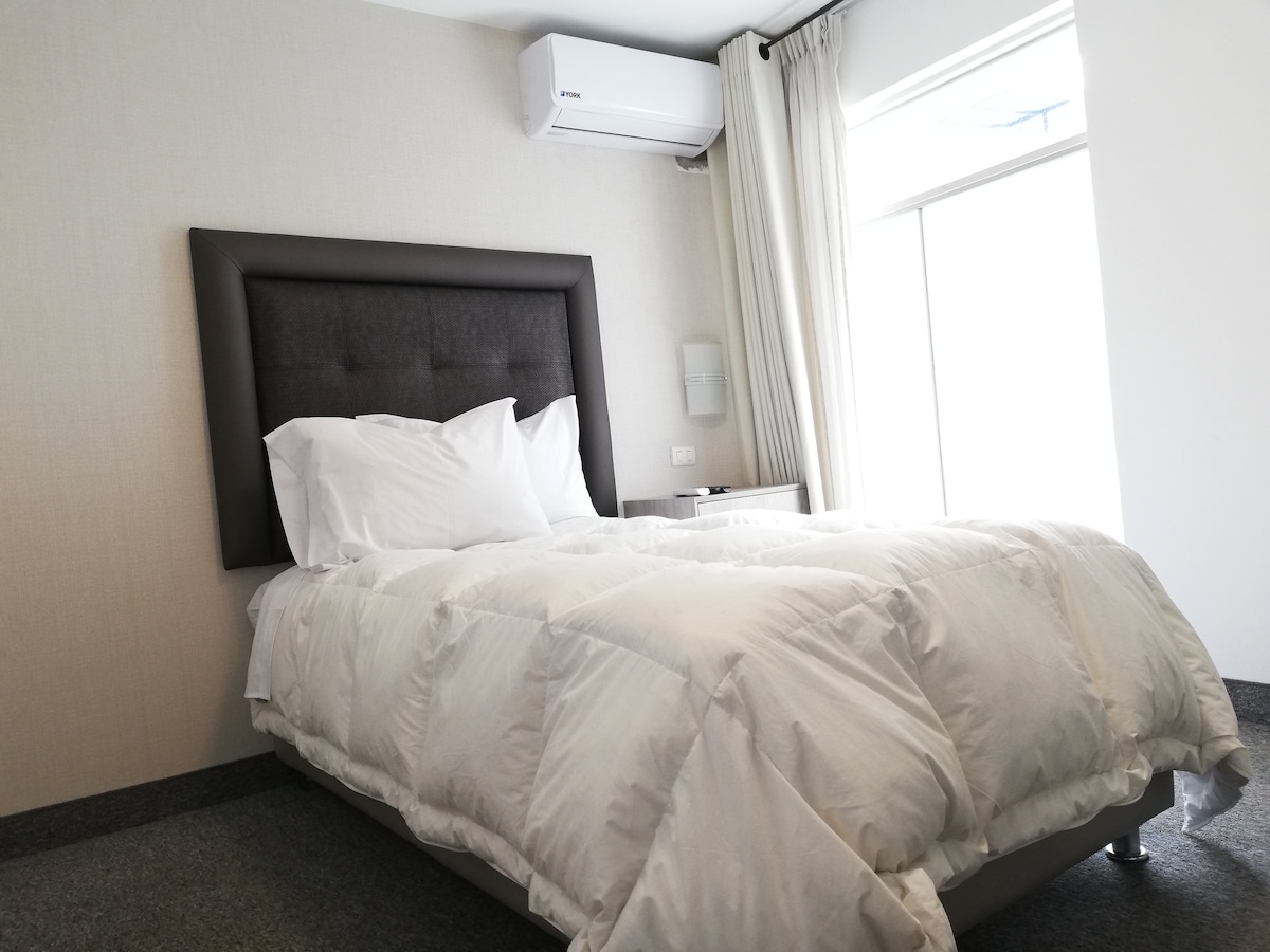 A spacious bed with white bedding is positioned against a light-colored wall, complemented by a dark upholstered headboard. Natural light enters through a large window with sheer curtains, illuminating the room's minimalistic design.
