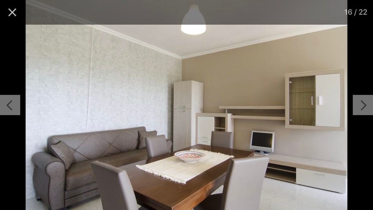 A contemporary living space features a brown sofa with geometric patterns, positioned next to a light wooden dining table surrounded by four grey chairs. A small TV sits on a sleek cabinet, and a bowl of decorative items rests on the table, adding a touch of style.