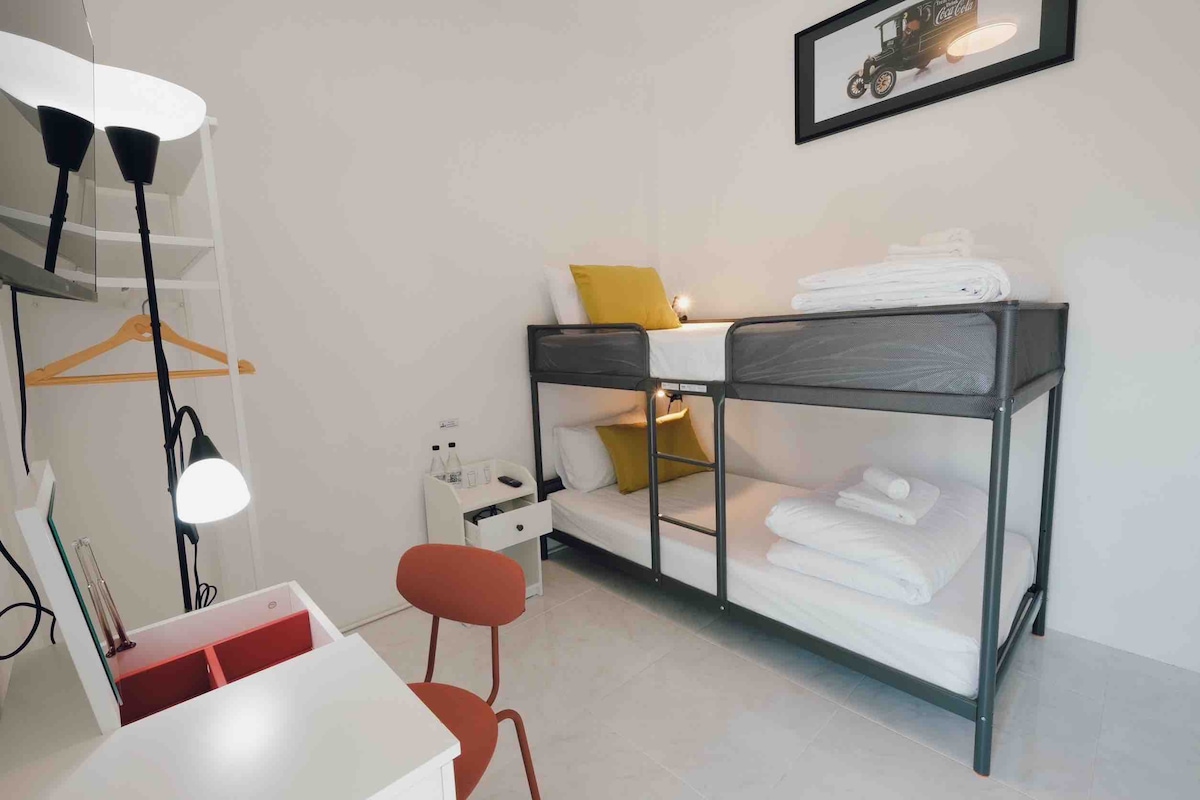 A bright sleeping area is furnished with a bunk bed, featuring neatly arranged linens and pillows. A desk with a chair and task lighting is positioned adjacent to the bed. Minimalist shelving holds essentials, while a framed artwork adorns the wall above.