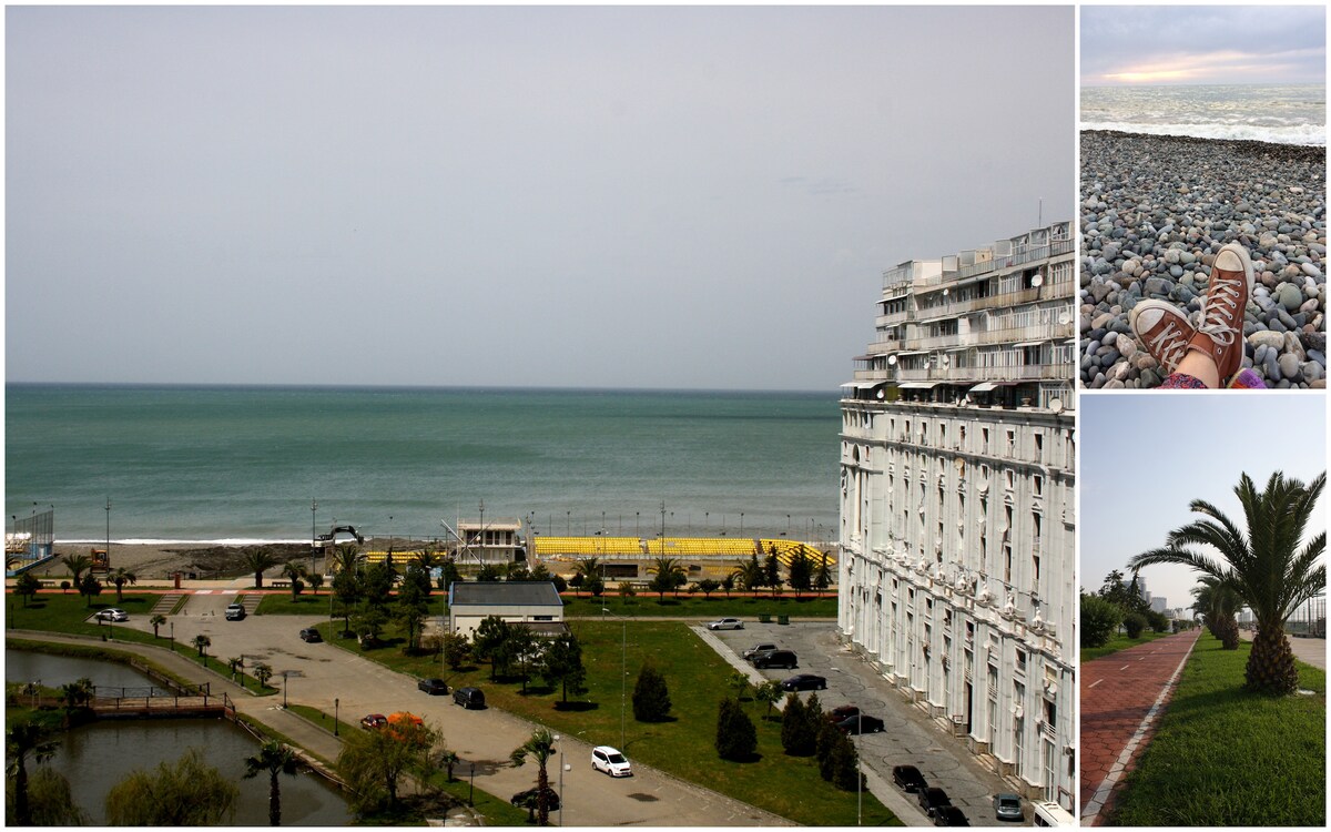 A panoramic view of the coastline reveals a tranquil sea with gentle waves lapping at the shore. In the foreground, a spacious area features palm trees and a landscaped path, with parked cars visible nearby. A beach area stretches along the waterfront, providing access to the sandy shore.