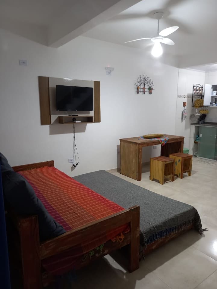 sala de estar con sofá cama con cocina americana, ventilador de techo, televisión inteligente de32pulgadas y mesa con 4 taburetes.