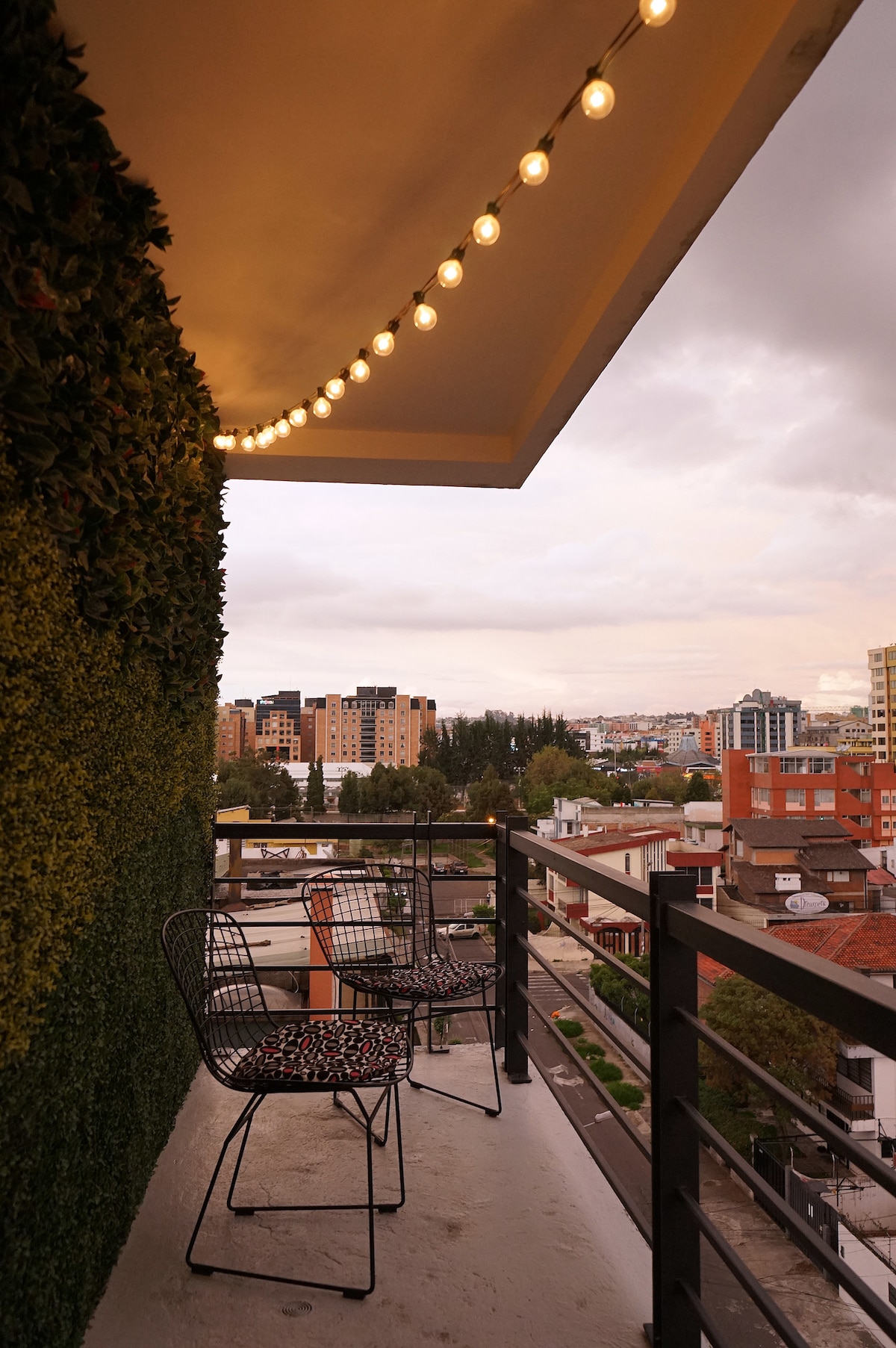 A balcony is displayed with two wire chairs positioned against a green wall, adorned with string lights overhead. The view includes city buildings and a skyline under a cloudy sky, showcasing a blend of urban scenery.