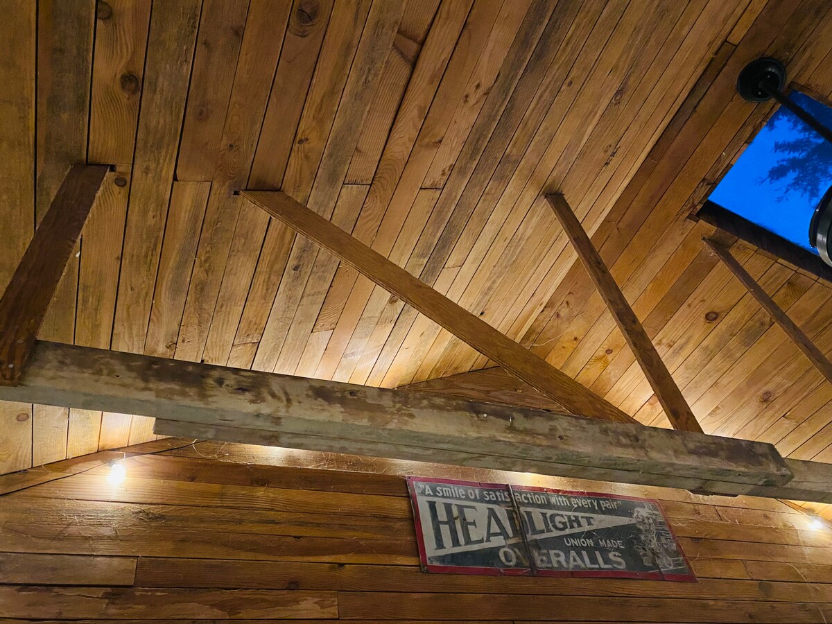 The image captures a close-up view of a wooden ceiling featuring exposed beams and a rustic aesthetic. Soft lighting highlights the natural grain of the wood, while a vintage sign is mounted on the wall below, contributing to the overall industrial decor of the space.