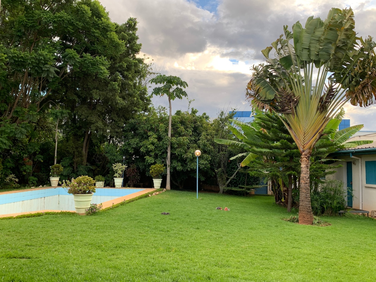 A lush green garden is showcased, featuring well-maintained grass and tropical plants. A swimming pool is visible, framed by white planters. Nearby, a house is partially seen, with blue accents against the natural backdrop. The sky is partly cloudy, adding dimension to the serene setting.