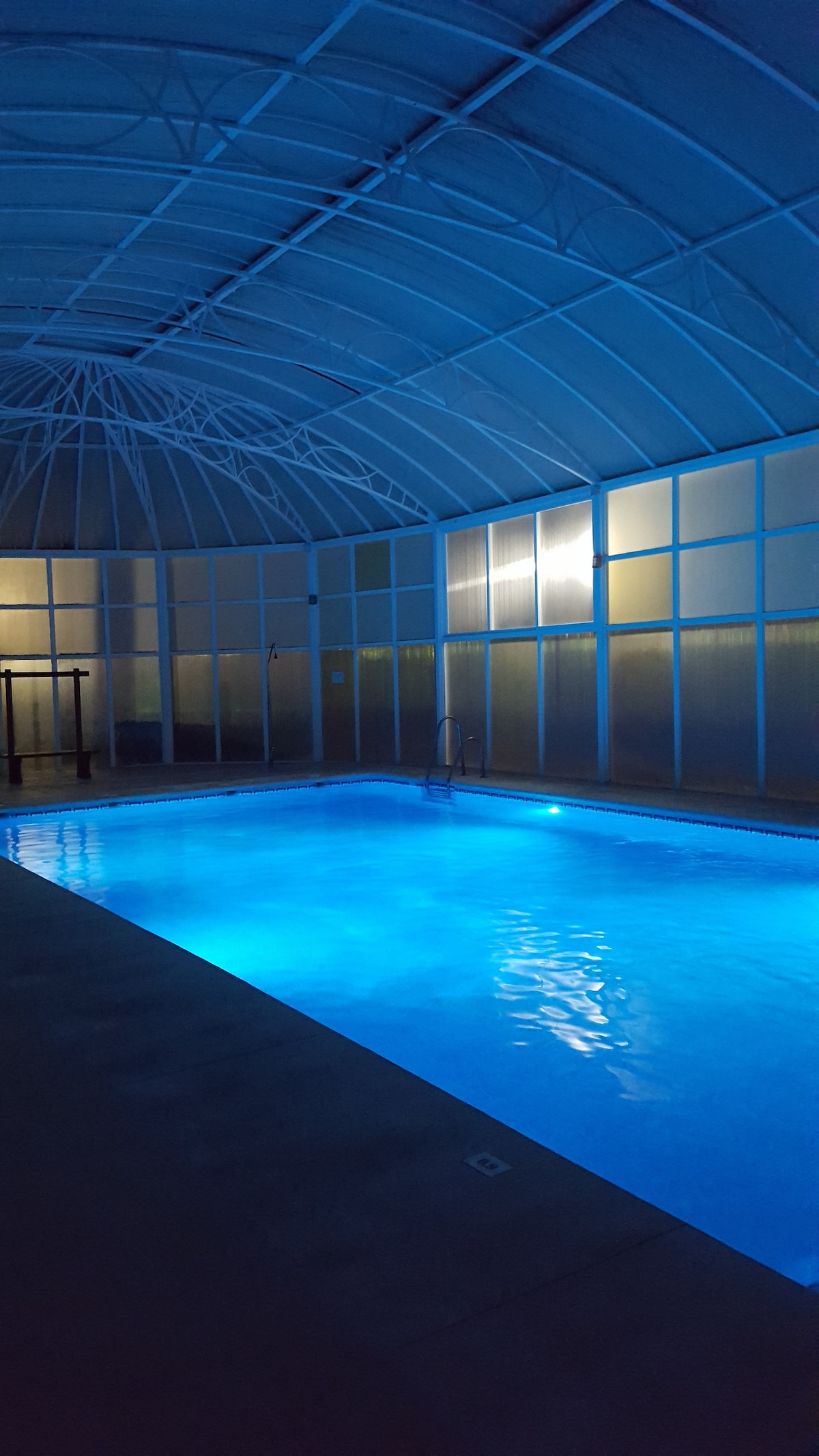 An indoor pool is illuminated by soft blue lighting, creating a calming atmosphere. The high ceiling showcases a supportive structure, with large windows allowing natural light to softly filter in. Water reflects the light, enhancing the serene environment.