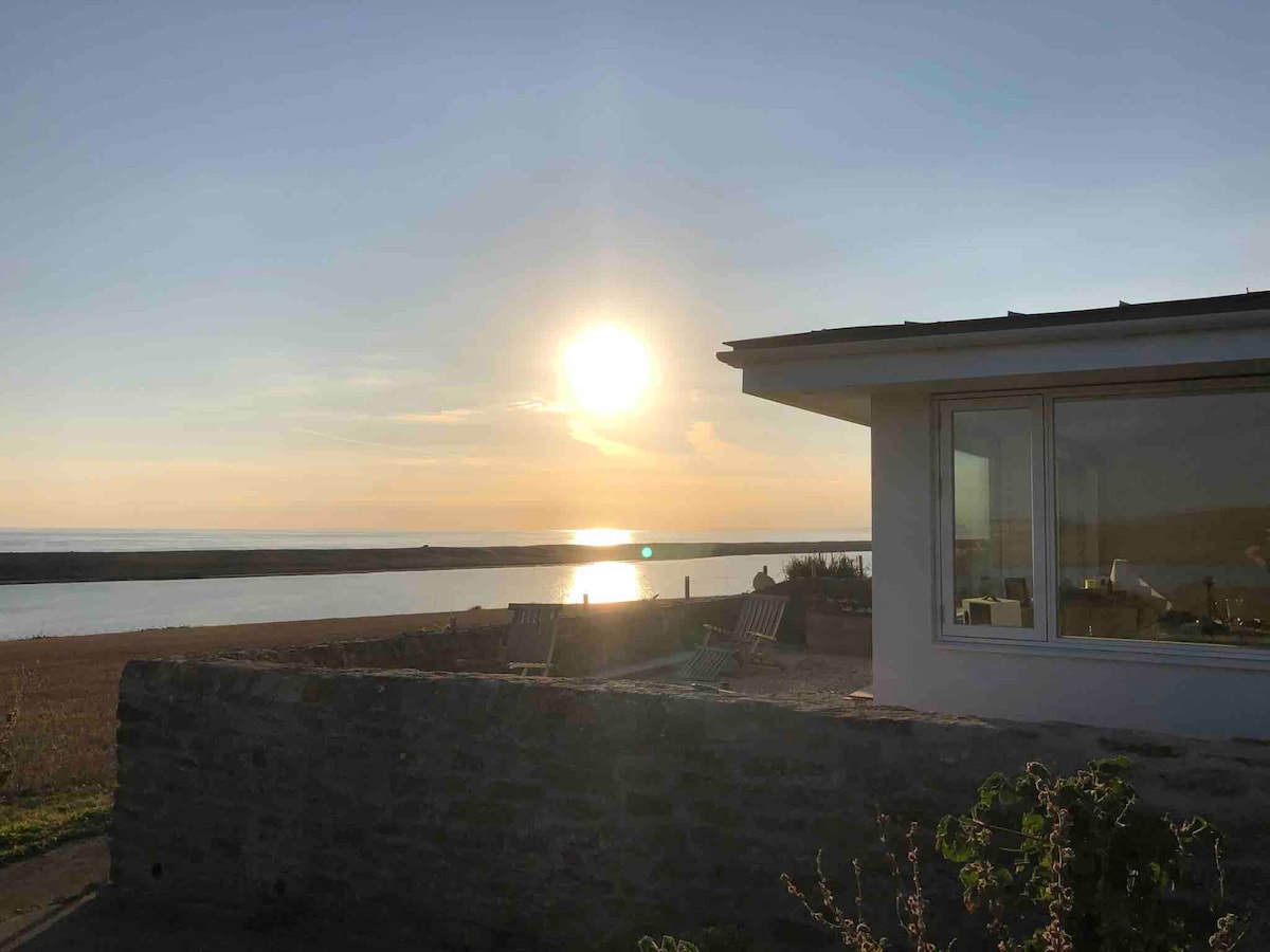 A spacious corner of a modern property is framed by a stone wall, overlooking a serene coastal scene. The sun sets on the horizon, casting warm reflections on the calm water. Comfortable seating can be seen in the foreground, inviting relaxation.