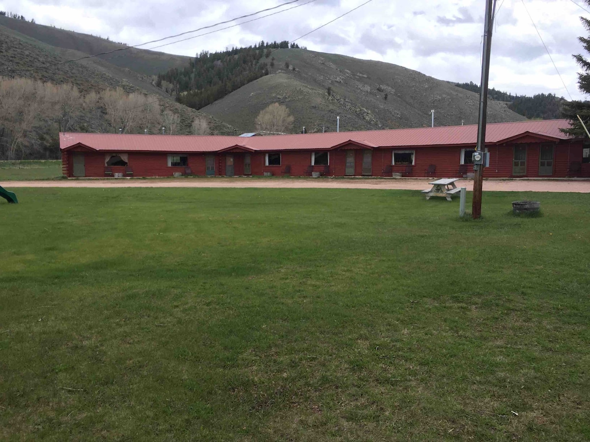 A rustic cabin exterior is presented, featuring a red facade and a metal roof. The building is flanked by grassy areas and set against a backdrop of rolling hills. A picnic table and a fire pit occupy the lawn, enhancing the cabin’s outdoor space.
