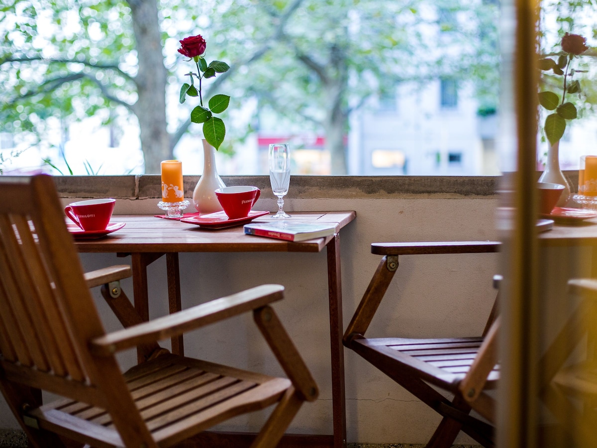 A cozy outdoor balcony setting is displayed, featuring a small wooden table with two chairs. A single red rose in a vase stands alongside a candle and a glass. Soft greenery is visible in the background, creating a pleasant and inviting atmosphere.