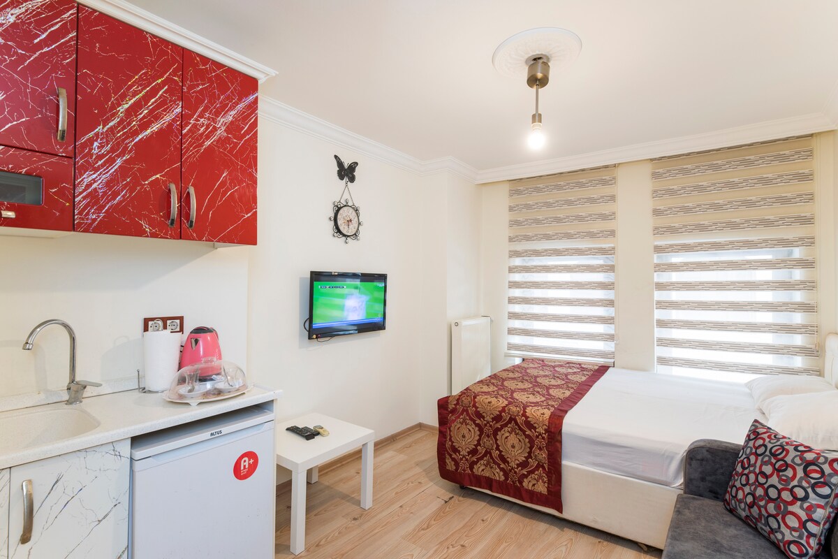A compact living space features a small kitchenette with red cabinetry and a mini refrigerator. A spacious bed with white linens is positioned next to a comfortable sofa. Natural light filters through patterned window shades, and a wall-mounted television adds functionality to the room.