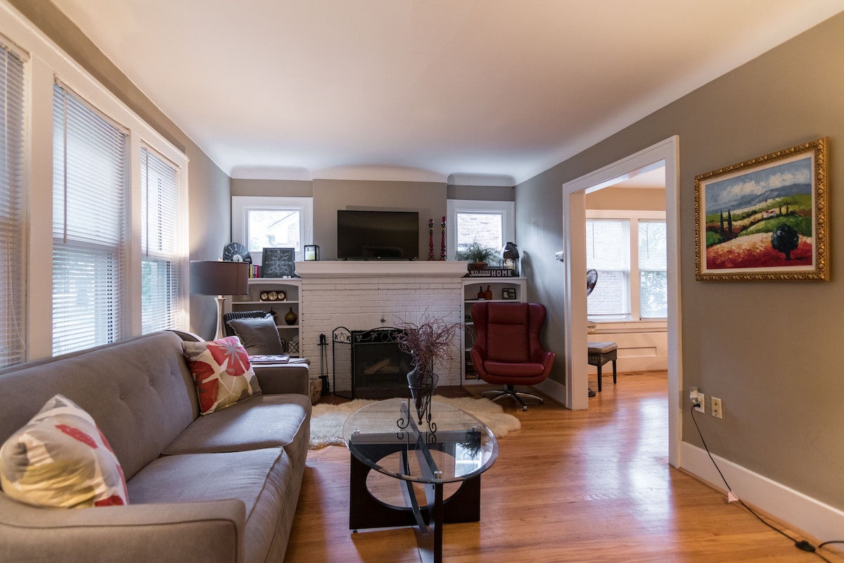 A living area features a comfortable sofa adorned with decorative pillows, positioned across from a minimalist coffee table. Natural light enters through multiple windows, illuminating the room. A fireplace is centrally located, complemented by a wall-mounted TV and a cozy armchair nearby.