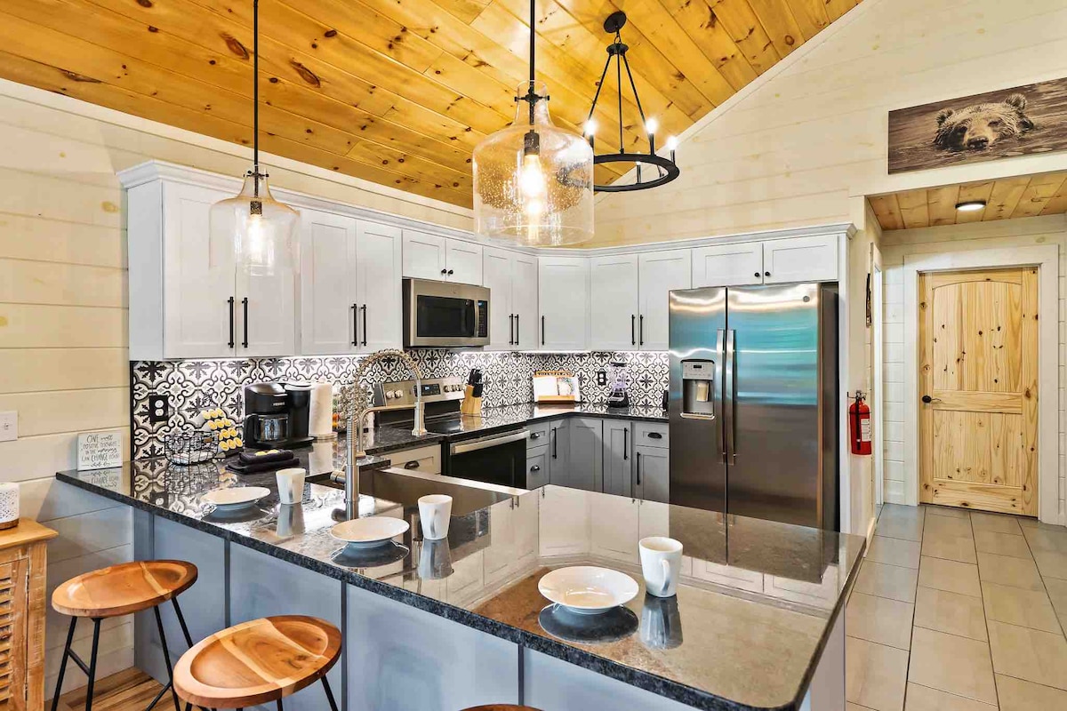 The kitchen features modern stainless steel appliances and an island with seating. A blend of light and dark cabinetry is complemented by bold patterned backsplash tiles. Warm wooden accents are visible in the ceiling and decor, contributing to a cozy atmosphere.